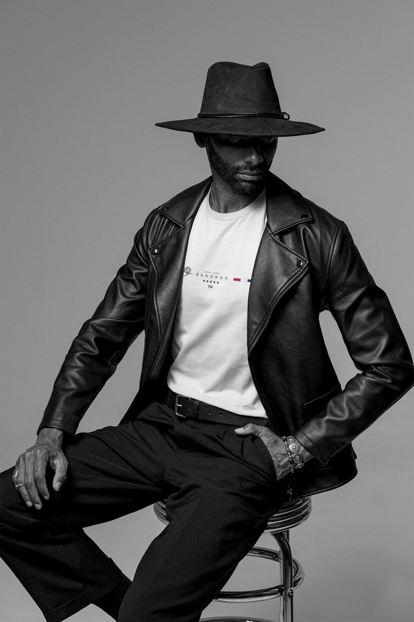 Seated male model wearing a white Krasswerk “BANGKOK” t-shirt under a black leather jacket. The model sits on a metallic stool, facing slightly to the side with his head tilted downward under a wide-brimmed hat. The shirt features a minimalist chest design with a tree icon, the word "BANGKOK," star details, the TH country code, and the Thai flag bar. Shot in studio with a smooth grey backdrop and dramatic black-and-white contrast.