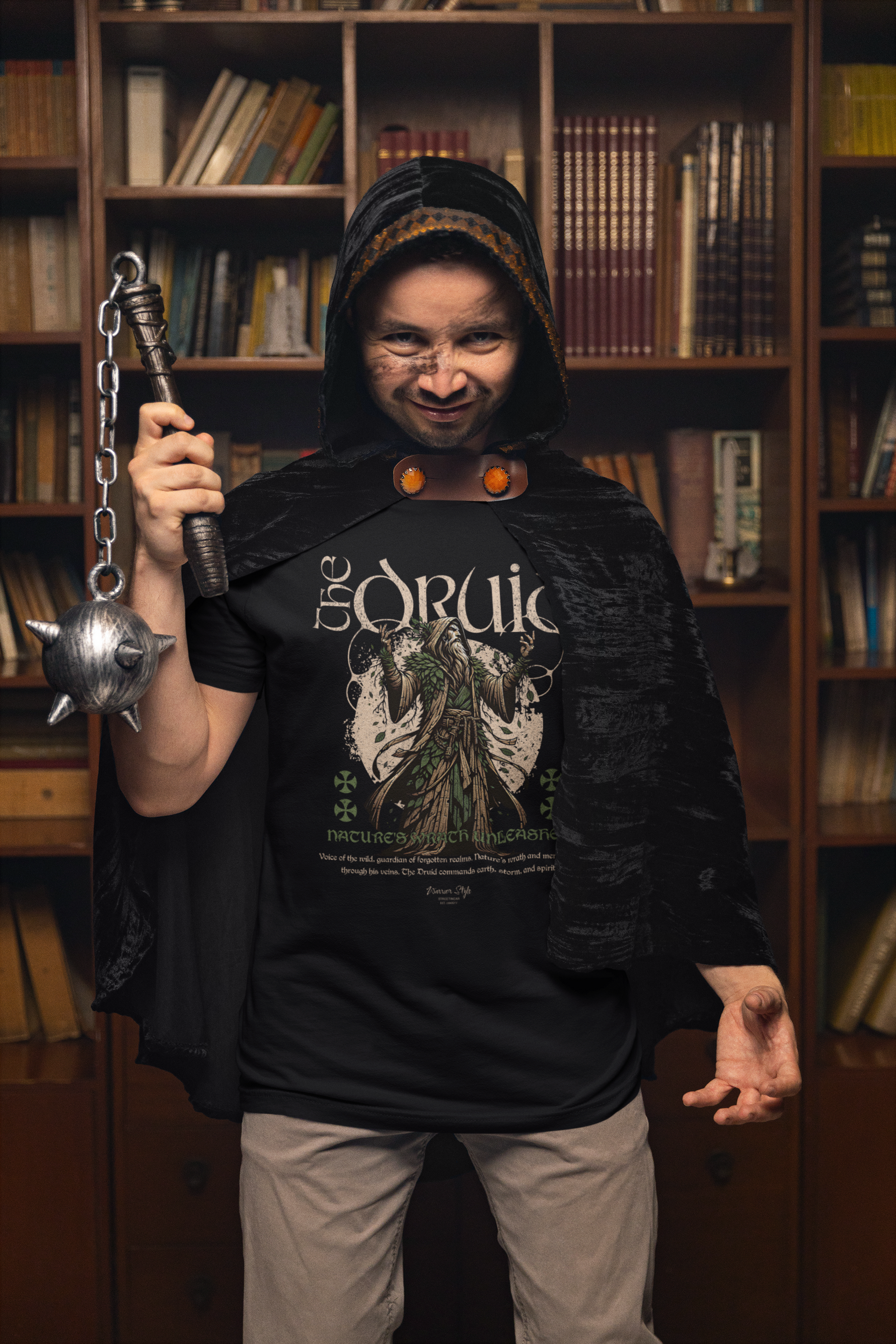 Male model in a fantasy setting dressed as a druid with hood and mace, standing in a library while wearing “The Druid” t-shirt. The mystical design and forest-green robe illustration are clearly visible.