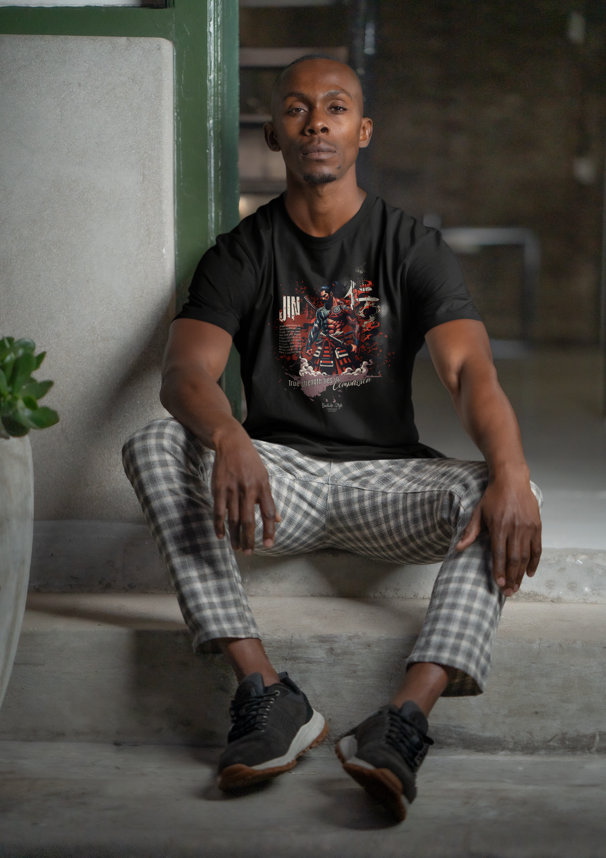 “JIN” black tee by KRASSWERK – worn by man seated on concrete stairs, calm expression, indoor urban vibe, graphic highlights strength through compassion