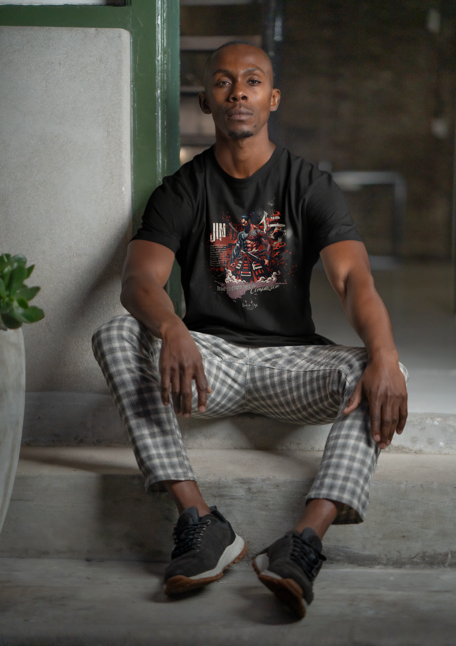 “JIN” black tee by KRASSWERK – worn by man seated on concrete stairs, calm expression, indoor urban vibe, graphic highlights strength through compassion