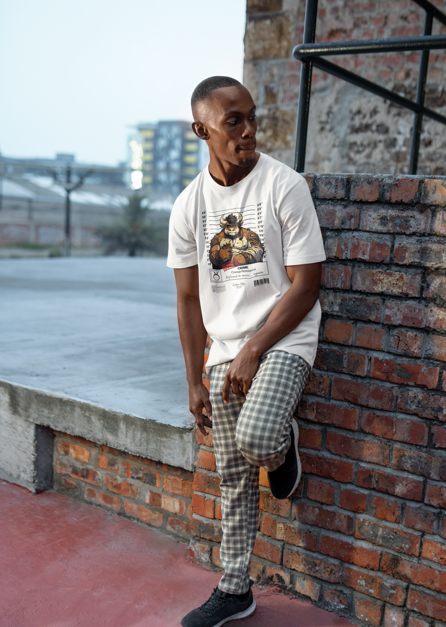 Urban outdoor scene — man leans against a brick wall wearing the TAURUS T-shirt in white. The shirt features a muscular bull in a police lineup, arms crossed, exuding quiet intensity. Text below reads “CRIME: Overprotective. Push him, regret it.” A bold visual for the unshakable types.