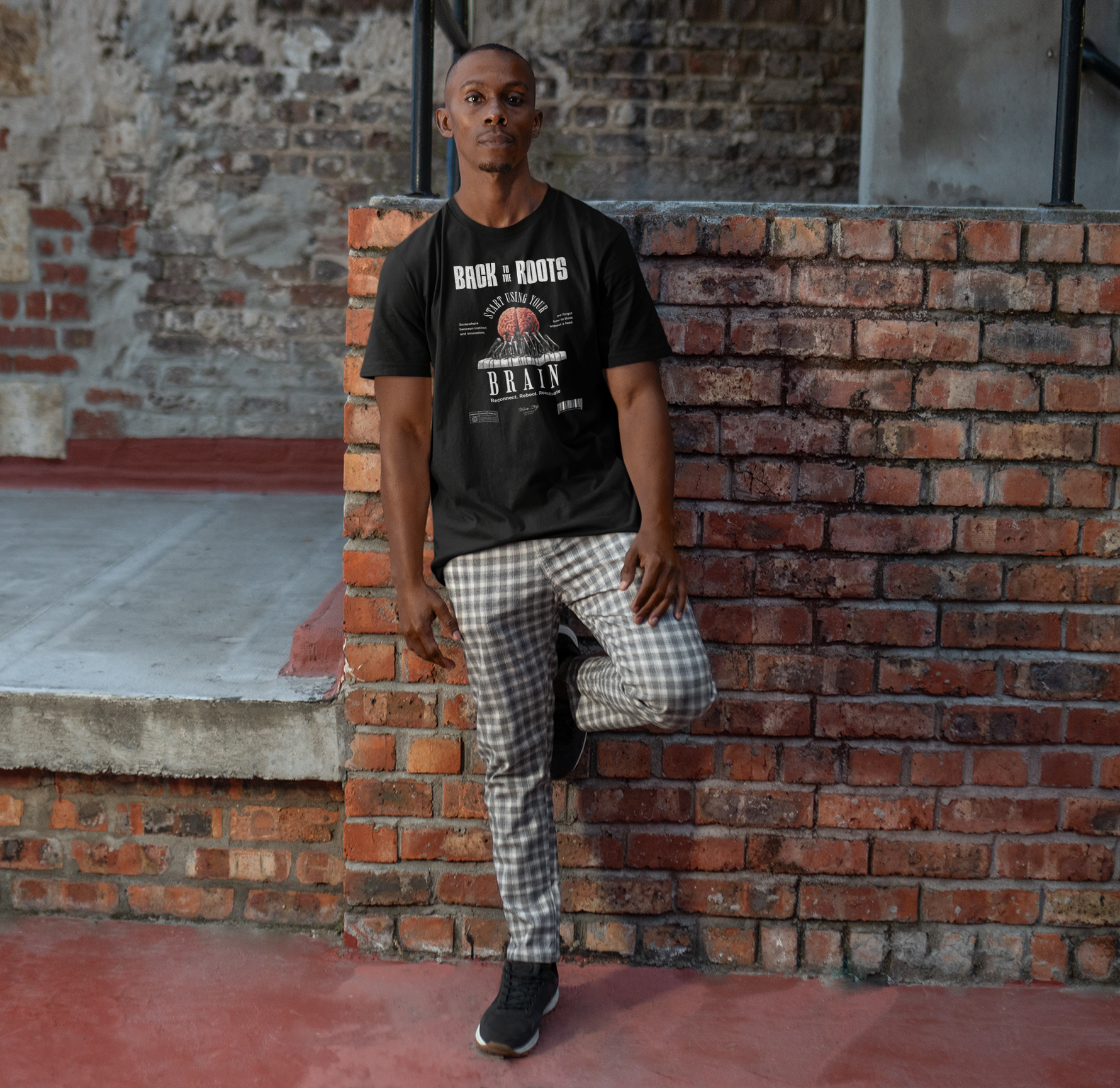 Male model leaning casually against a brick wall in a black “Back to the Roots” t-shirt, featuring a detailed brain and root design. Urban setting, relaxed stance, natural daylight highlights the message-driven graphic.