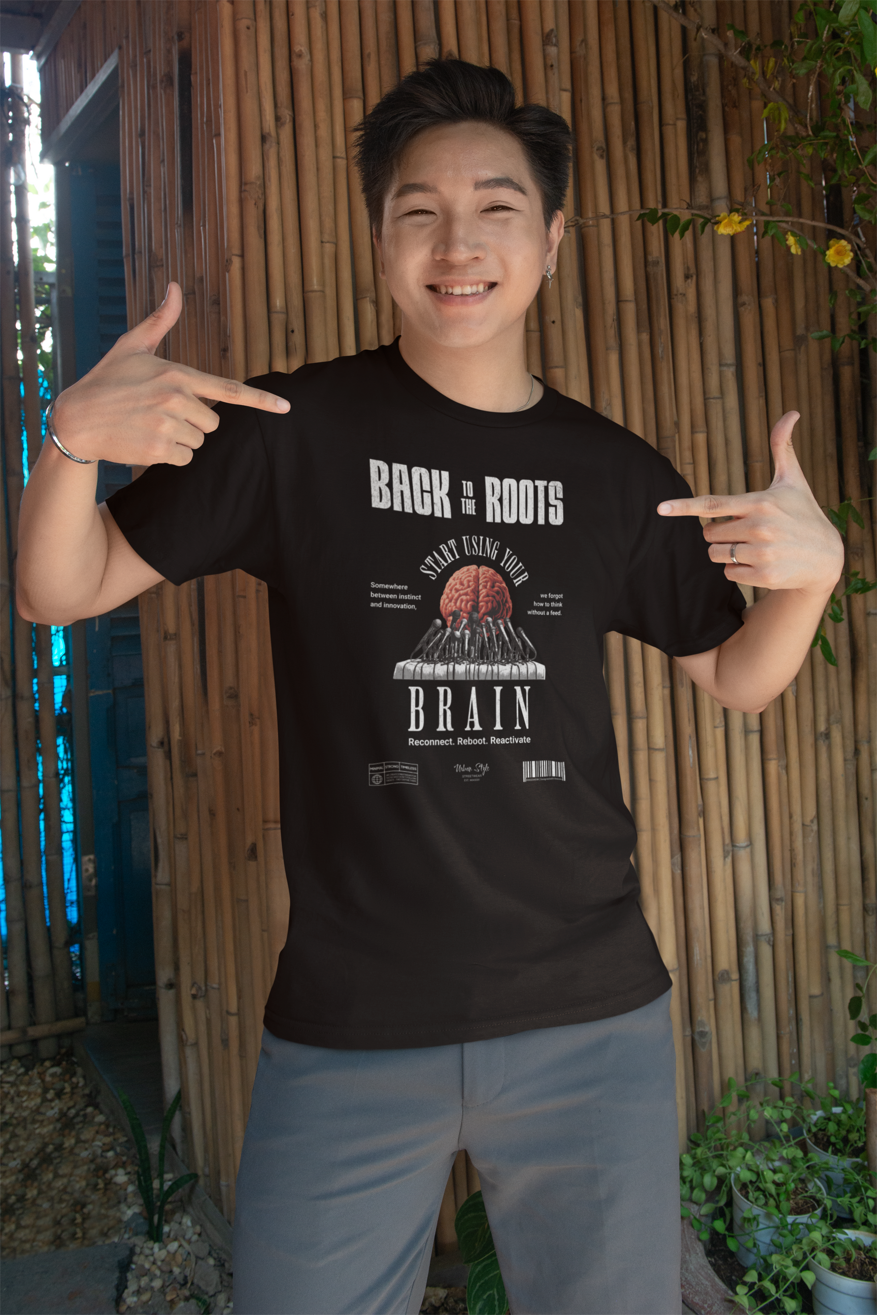Male model smiling and pointing at his black “Back to the Roots” t-shirt featuring a stylized brain. Natural outdoor setting with bamboo background and casual vibe.