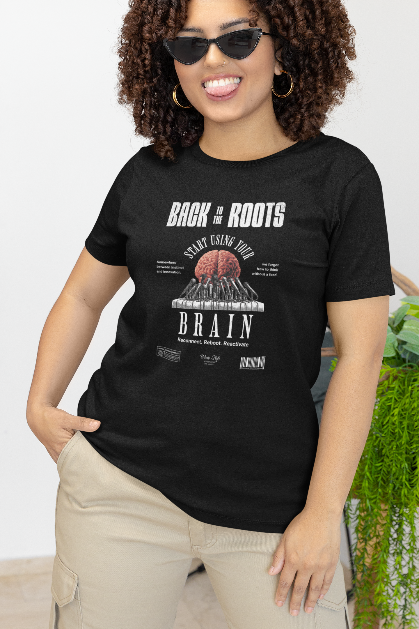 Female model smiling playfully in a black “Back to the Roots” t-shirt, featuring a stylized brain graphic. Indoor setting with plants, casual yet bold attitude matches the design’s message.