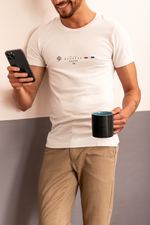 Smiling male model in a white Krasswerk “BANGKOK” t-shirt, holding a black ceramic mug in one hand and a smartphone in the other. The design on the shirt features the word Bangkok, a lotus flower icon, five stars, country code “TH,” and a red-blue horizontal flag bar. The background is softly lit in warm beige tones, creating a relaxed, casual morning vibe.