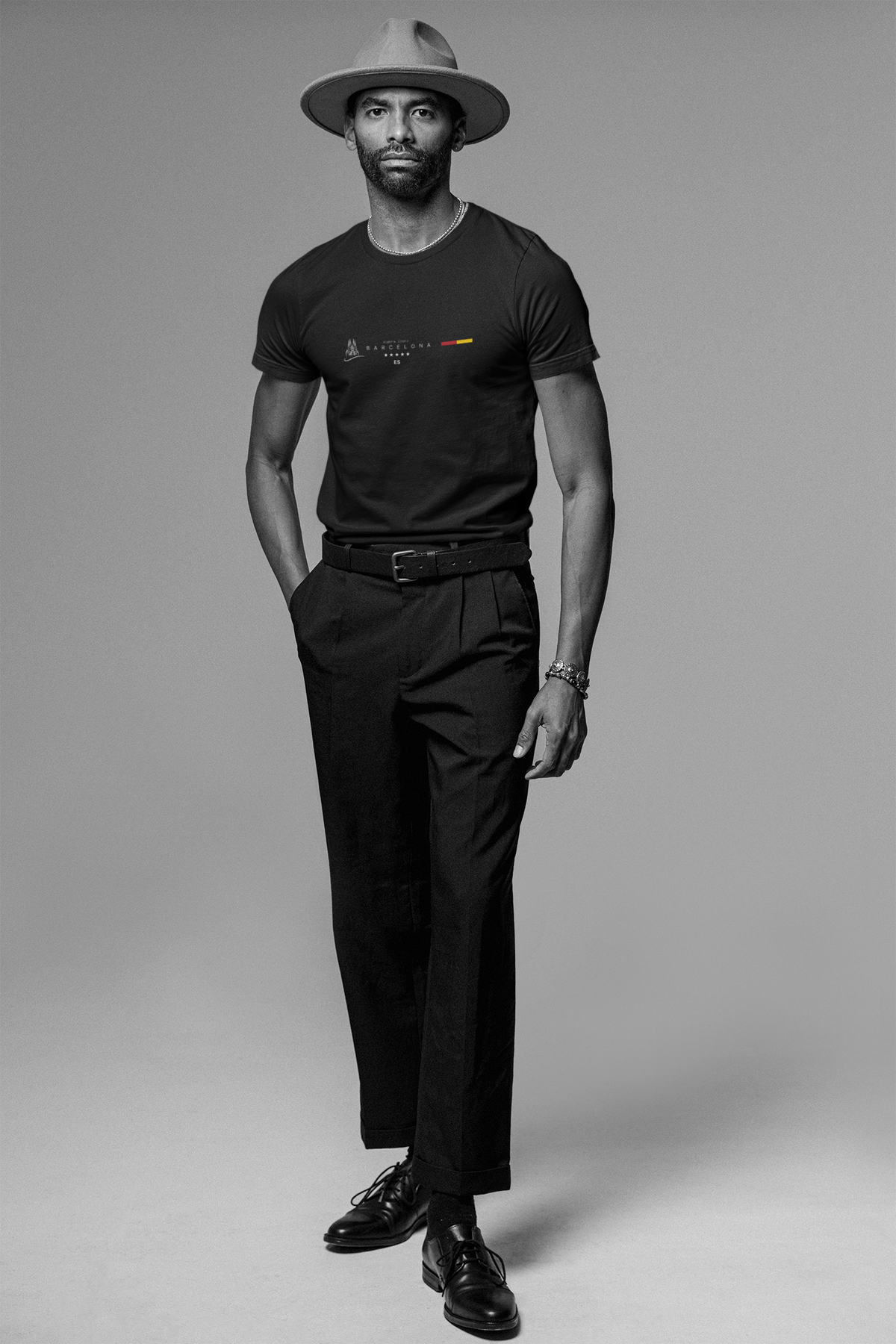 A tall, stylish man poses confidently in a black “BARCELONA” Krasswerk t-shirt with the Spanish flag and minimal typography centered on the chest. He wears tailored black pants, polished leather shoes, and a wide-brimmed hat. The monochrome setting adds sophistication and contrast, emphasizing the shirt’s sharp graphic design.
