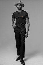 A tall, stylish man poses confidently in a black “BARCELONA” Krasswerk t-shirt with the Spanish flag and minimal typography centered on the chest. He wears tailored black pants, polished leather shoes, and a wide-brimmed hat. The monochrome setting adds sophistication and contrast, emphasizing the shirt’s sharp graphic design.