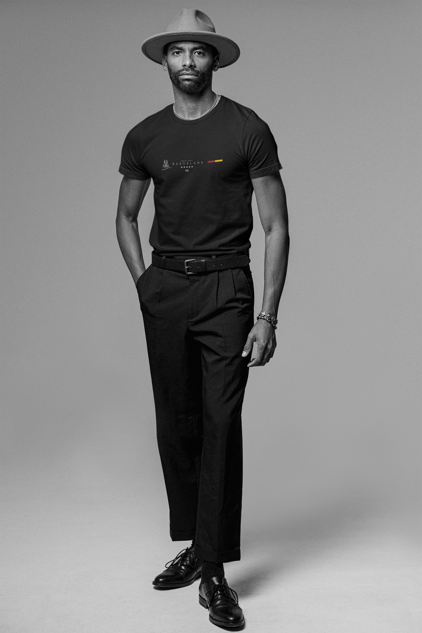 A tall, stylish man poses confidently in a black “BARCELONA” Krasswerk t-shirt with the Spanish flag and minimal typography centered on the chest. He wears tailored black pants, polished leather shoes, and a wide-brimmed hat. The monochrome setting adds sophistication and contrast, emphasizing the shirt’s sharp graphic design.