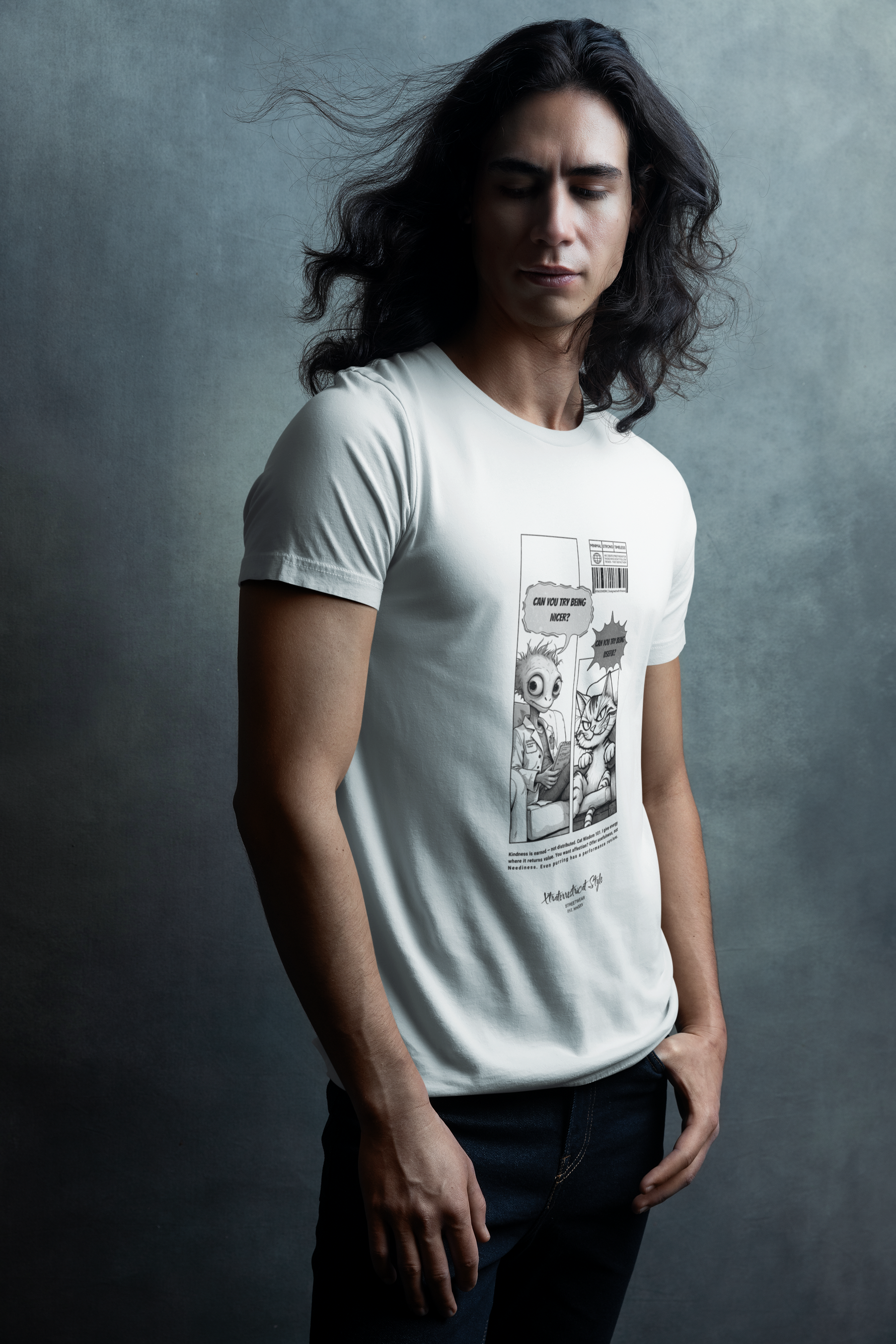 A man with long wavy hair poses in front of a textured gray wall, wearing a white graphic t-shirt and dark jeans. The t-shirt features a comic-style illustration where an alien asks, “Can you try being nicer?” and a sarcastic cat responds, “Can you try being useful?” His calm expression and the soft lighting give the scene an artistic, editorial feel.