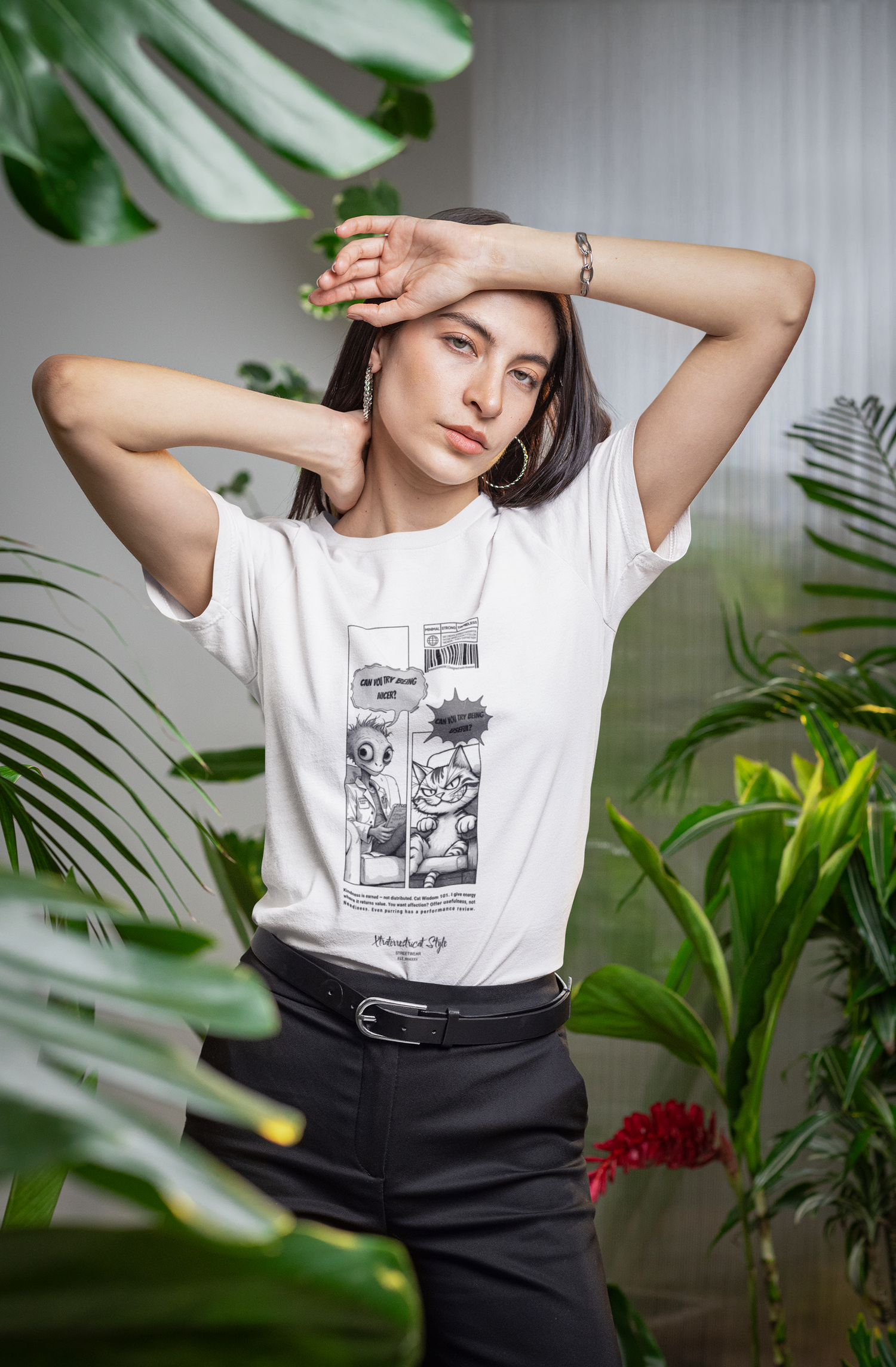 A stylish woman posing indoors among large green tropical plants, wearing a white graphic t-shirt tucked into high-waisted black pants. The shirt features a comic-style black-and-white illustration where an alien asks, “Can you try being nicer?” and a sarcastic cat replies, “Can you try being useful?” Her expression is confident and relaxed, with one arm raised over her forehead, adding to the artistic and natural vibe of the shot.