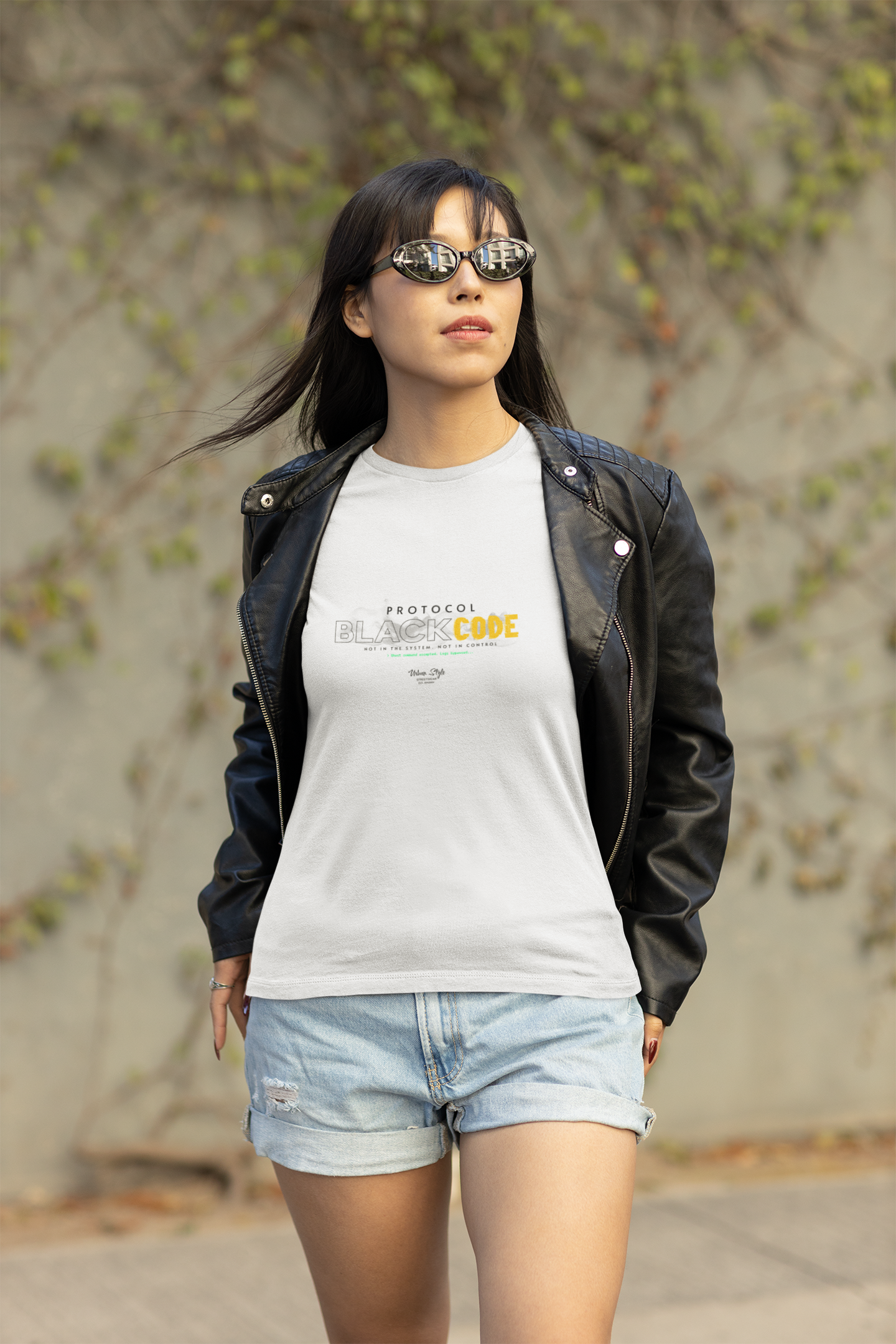 Outdoor lifestyle shot of a woman in sunglasses and a black leather jacket, walking confidently in daylight while wearing a white “PROTOCOL BLACKCODE” t-shirt. The design features a smokey “BLACK” text and yellow “CODE” with the phrase “NOT IN THE SYSTEM. NOT IN CONTROL.” Background includes a blurred concrete wall with scattered ivy.