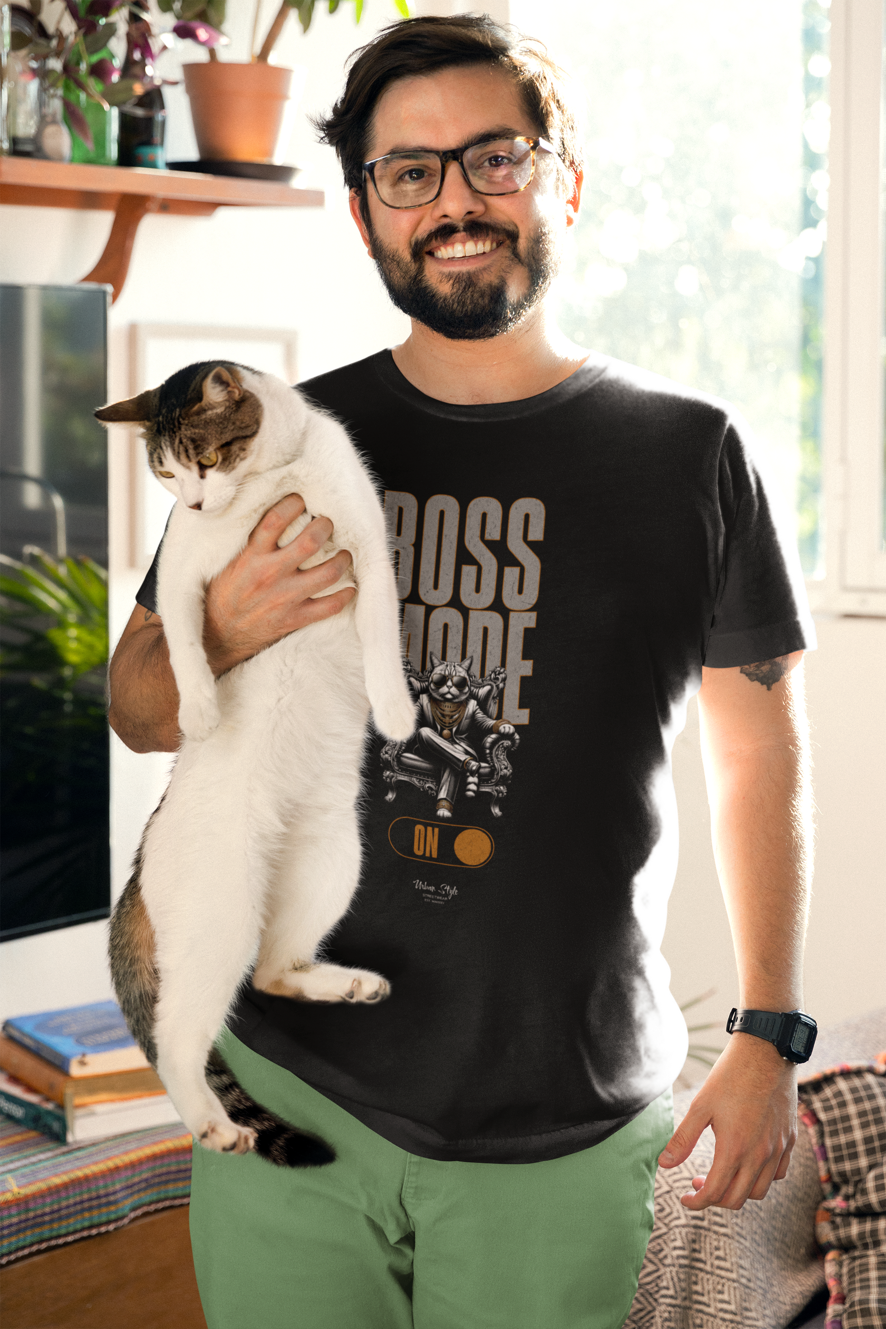 A cheerful male model holding a cat in a cozy, sunlit room, wearing the BOSS MODE T-shirt. The design features a cool character on a throne with a gold switch graphic labeled “ON.”