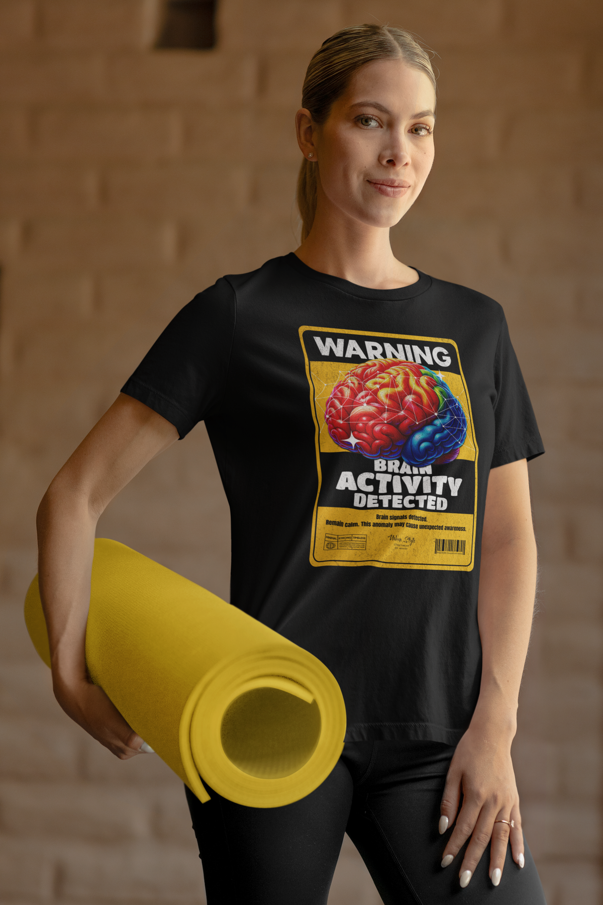 A sporty female model holding a rolled yellow yoga mat stands in a relaxed pose, wearing a black graphic t-shirt with a bold warning label: “WARNING – BRAIN ACTIVITY DETECTED.” The colorful 3D brain artwork contrasts sharply against the black tee, delivering a clever mix of humor and intellect.