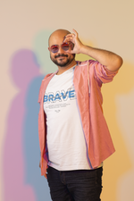 Man model wearing THE BRAVE t-shirt in white — smiling with confidence, holding up his fingers in a playful gesture over round sunglasses. The bold “BRAVE” print stands out on the shirt with a blue layered design and the phrase “Stay brave. Stay wild.” A pastel gradient background adds a fun, expressive vibe to the shot.