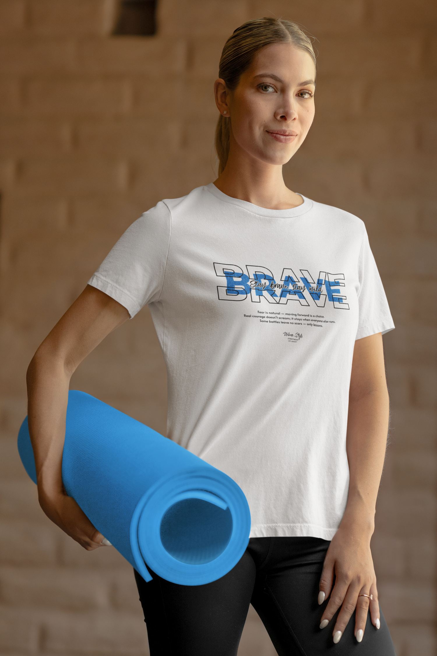 Woman model wearing THE BRAVE t-shirt in white — standing relaxed with a yoga mat in hand, soft smile and steady gaze. The layered “BRAVE” typography stands bold on the chest, with the phrase “Stay brave. Stay wild.” highlighted in soft blue. Clean indoor setting with warm tones keeps the focus on courage and calm.