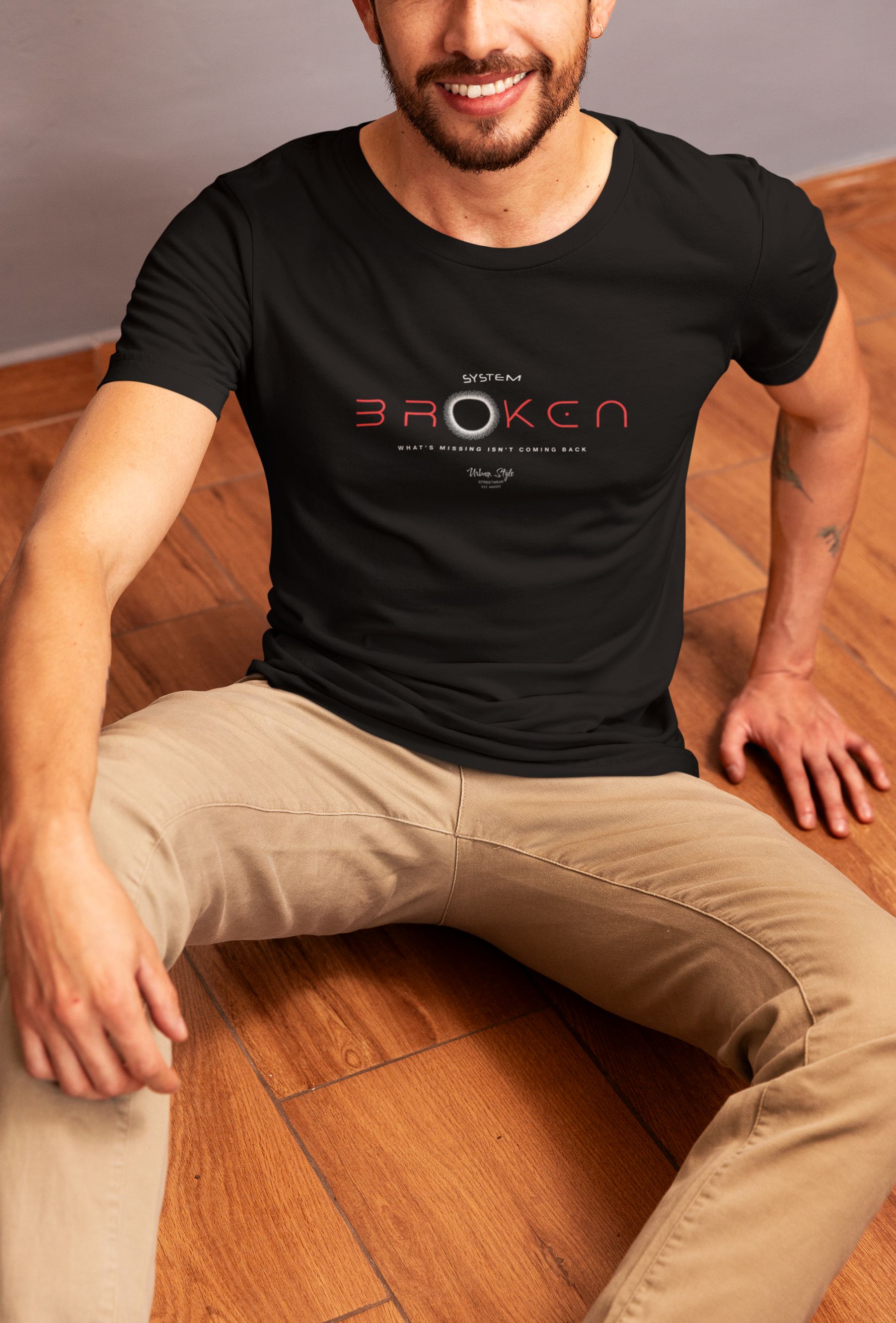 Man wearing a black t-shirt with a logo sitting on a wooden floor.