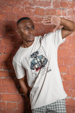 A male model poses in front of a rustic red brick wall, raising his hand as if shielding his eyes from light. He wears the white “CAPRICORN” tee featuring a horned businessman holding coffee and files. The urban background adds contrast to the clean, illustrative design.