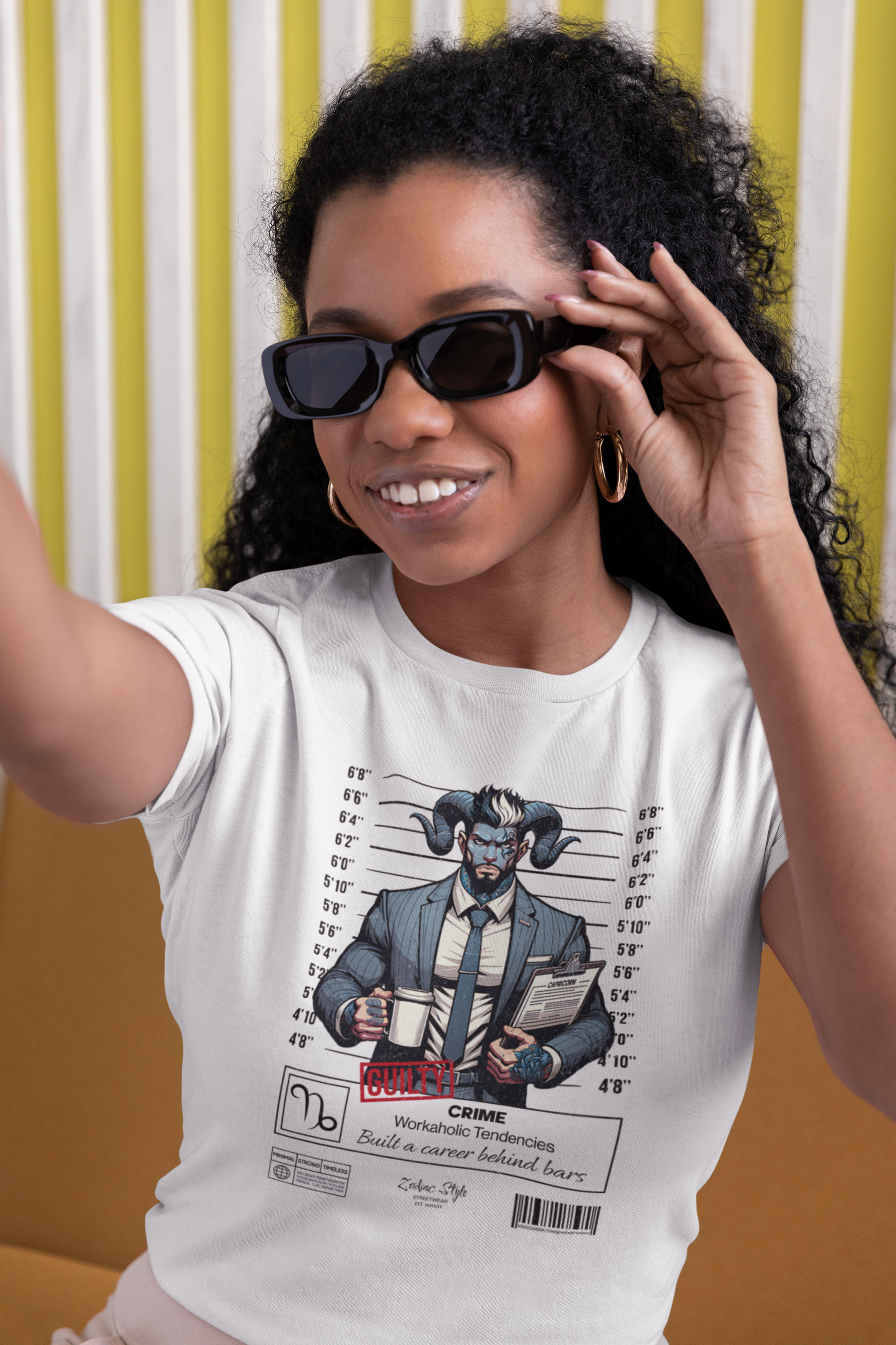 A cheerful female model with curly hair and sunglasses smiles while wearing the white “CAPRICORN” graphic tee. The design shows a horned man in a suit holding a mugshot board and coffee. Vertical yellow stripes in the background enhance the bright, casual vibe.
