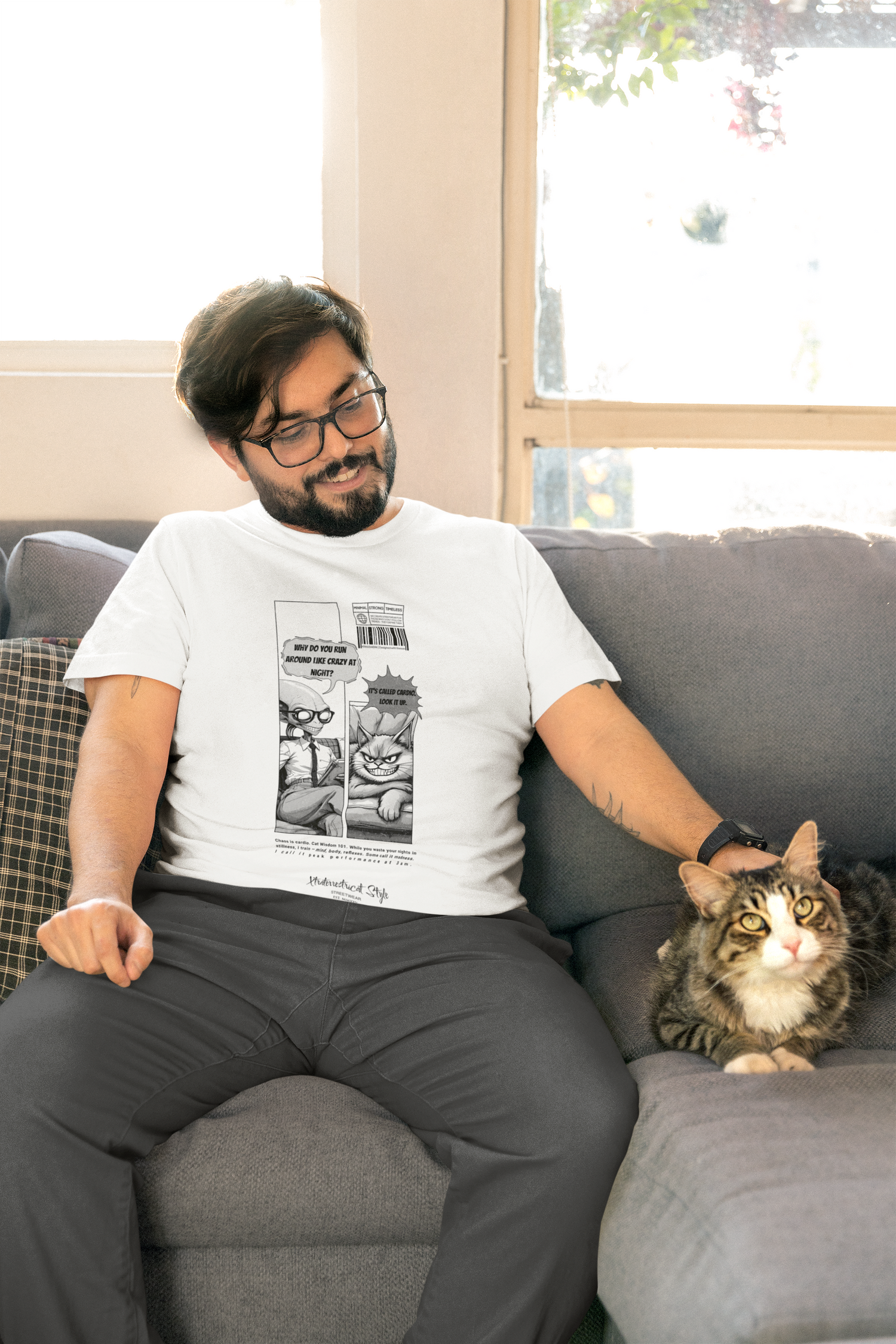 A man sits casually on a grey sofa next to a tabby cat, wearing a white t-shirt with a comic-style design. The design features two panels — the cat says “It’s called cardio. Look it up.” in response to the question “Why do you run around like crazy at night?” from another cat. Bright natural light enters through the window behind, giving the scene a cozy, playful vibe.