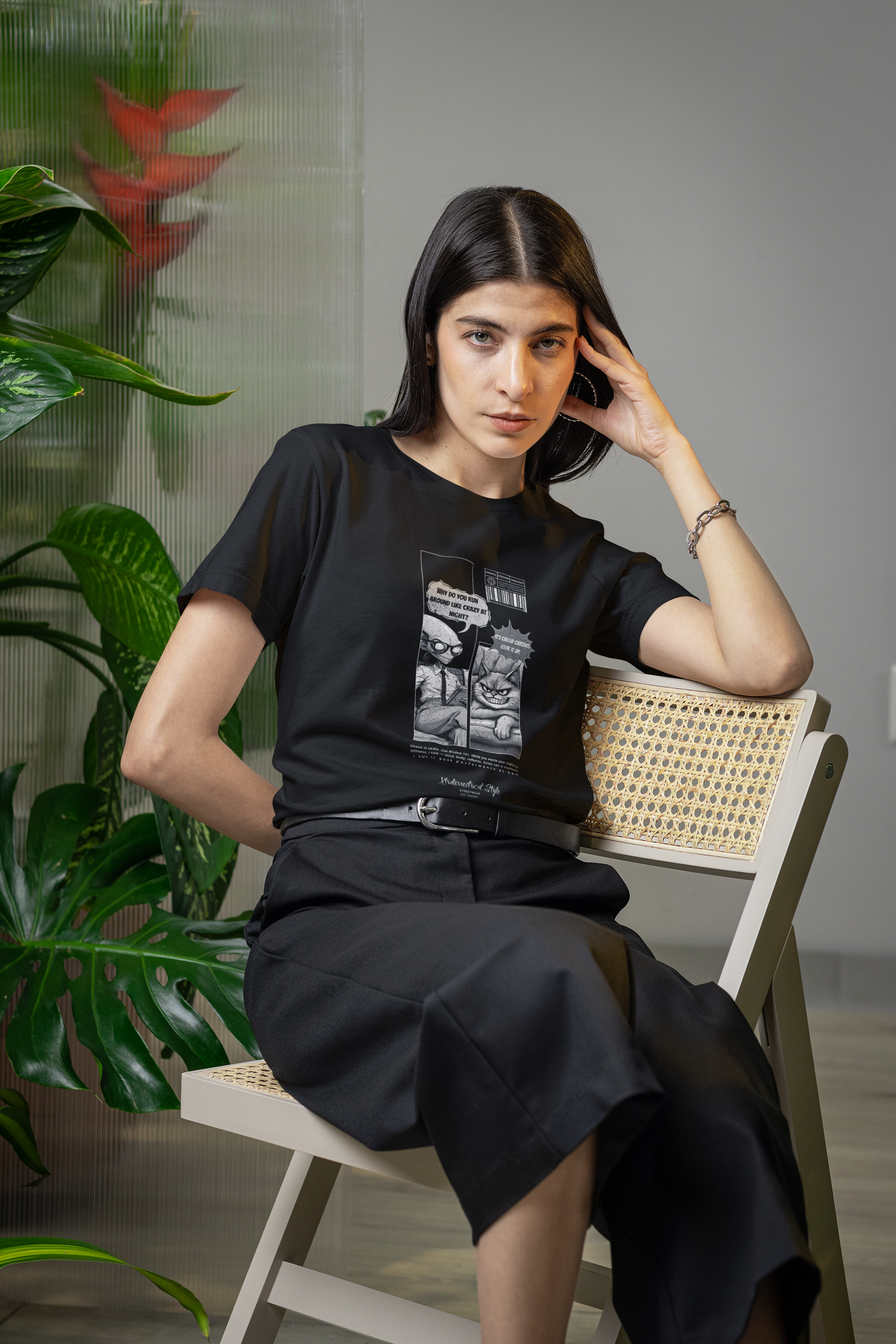 A woman sits on a woven-back chair wearing a black t-shirt with a black-and-white comic print. The design features an alien therapist and a sarcastic cat in a therapy session. The alien asks, “Why do you run around like crazy at night?” and the cat replies, “It’s called cardio. Look it up.” Large green leaves and a frosted glass panel with red flowers add a modern indoor jungle vibe to the scene.