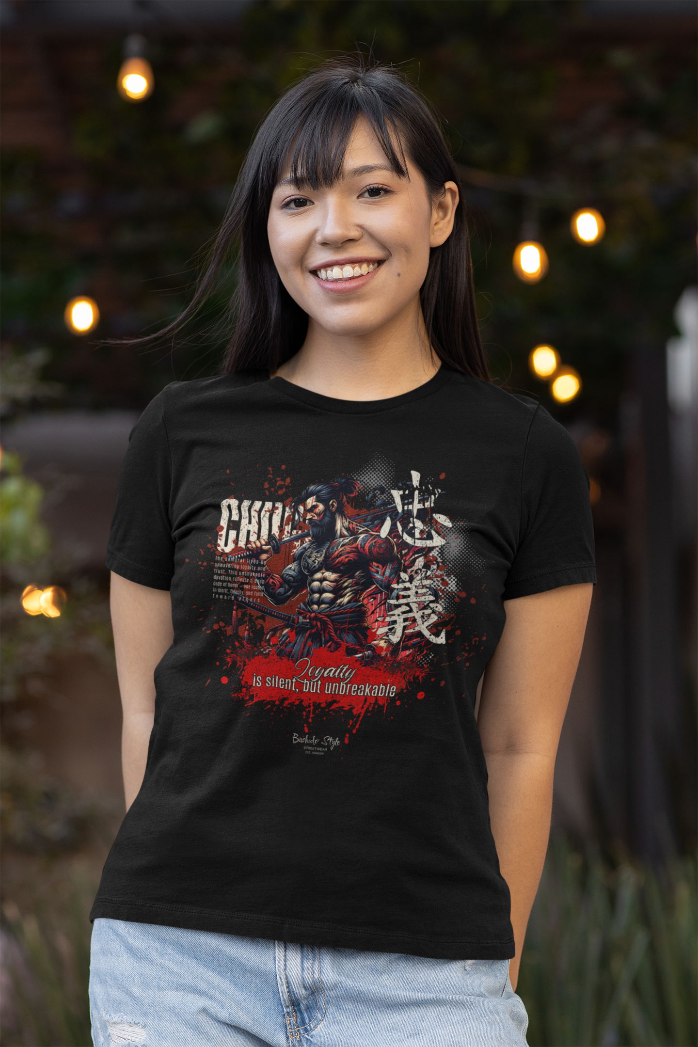 A smiling woman wearing the CHŪGI t-shirt from the 7 Virtues of Bushido series. The shirt features a fierce samurai with red and white ink splashes and the phrase "Loyalty is silent, but unbreakable." Soft outdoor lights glow in the background.