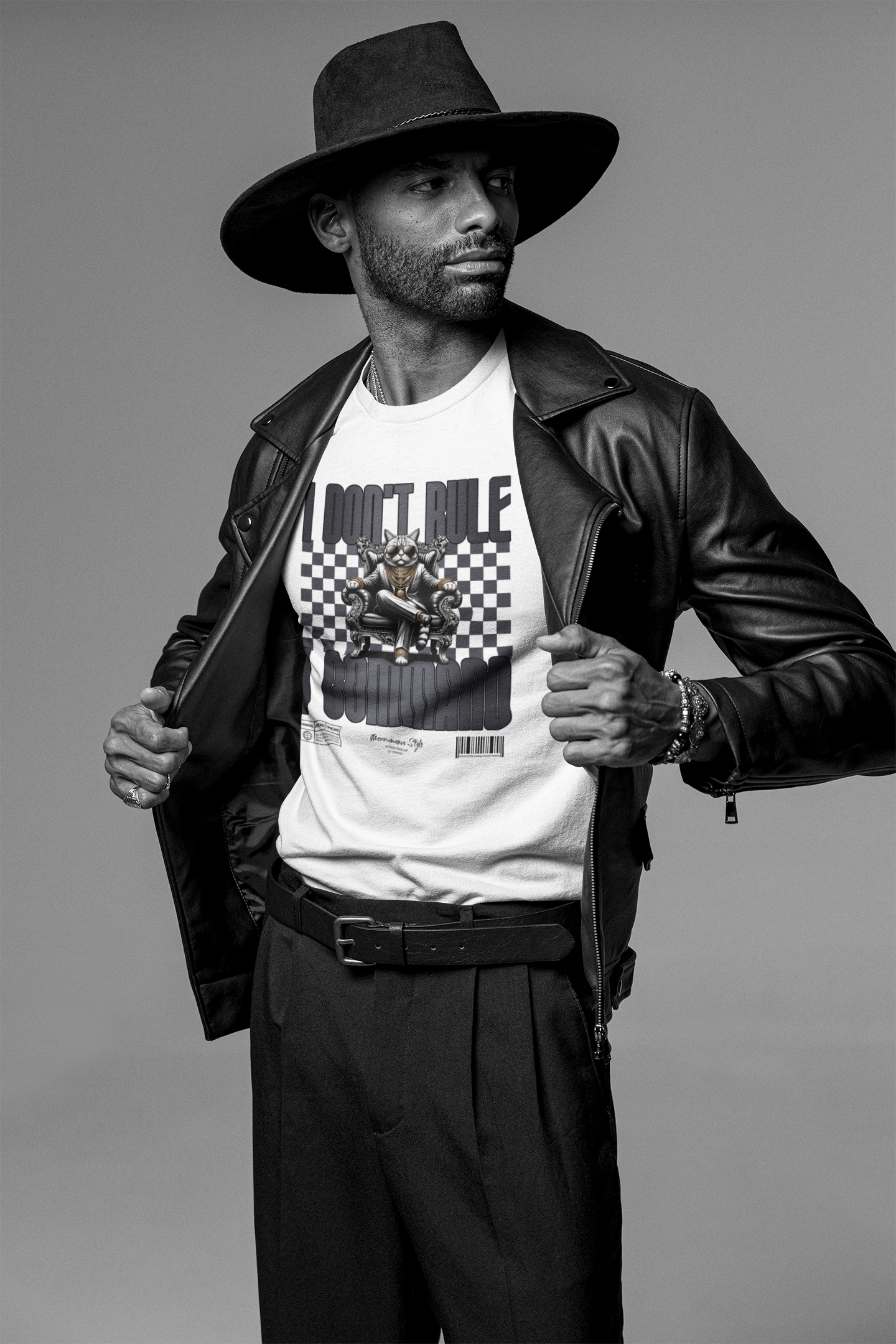 Man in black hat and leather jacket revealing a white graphic tee with a throne-sitting cartoon cat. The shirt reads "I DON'T RULE. I COMMAND." with a bold checkered backdrop.