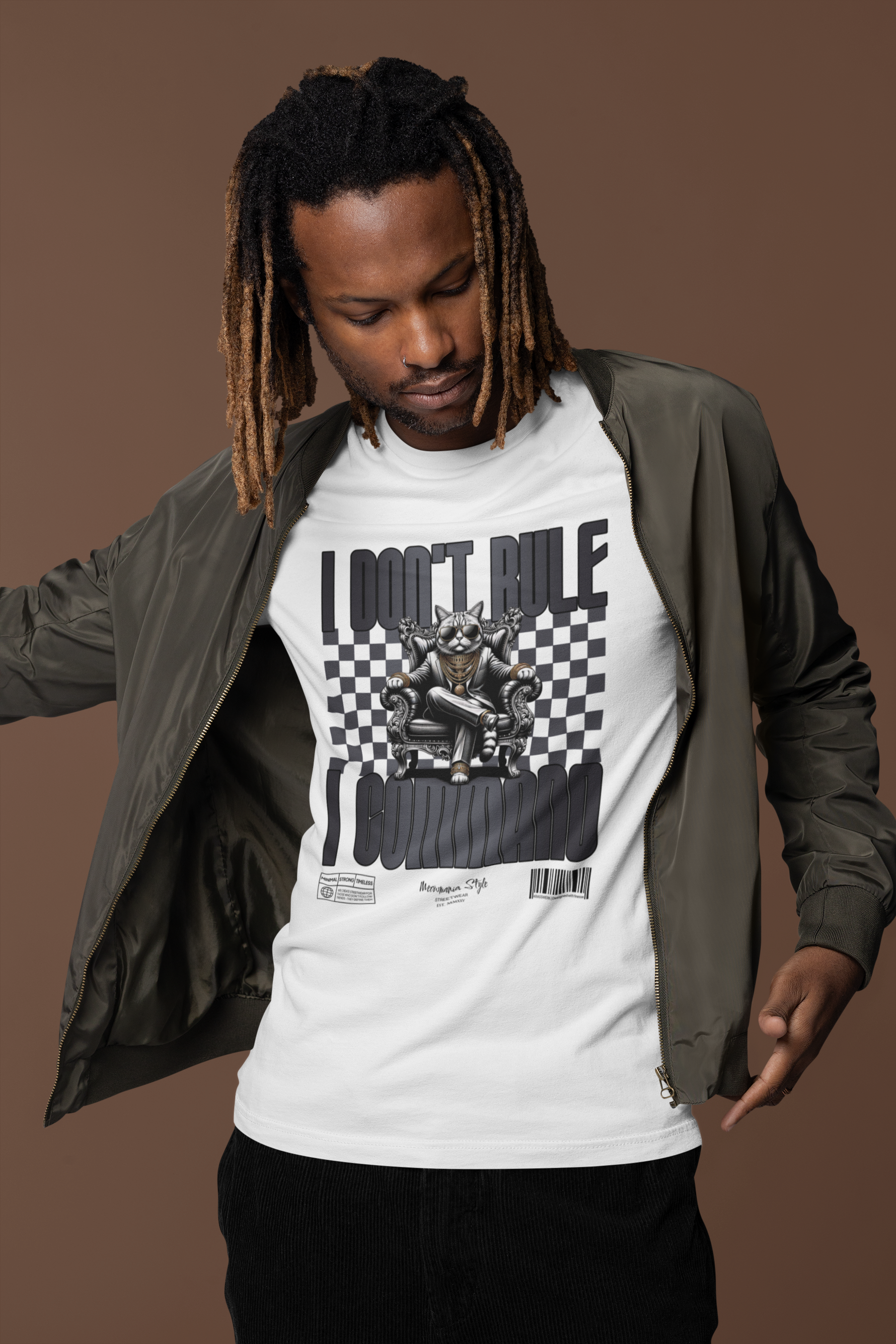 Man in a bomber jacket and corduroy pants pointing toward his white t-shirt, which features a commanding cat on a throne with checkered background and the phrase “I DON’T RULE. I COMMAND.” on a warm brown backdrop.