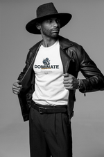 Man wearing a white DOMINATE t-shirt under a leather jacket, looking sideways with calm intensity. The bold lettering and mechanical fist design capture a message of controlled force — dominance without noise.