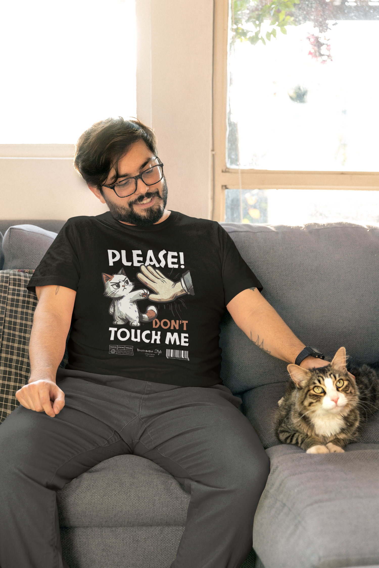 A smiling man sits on a sofa next to a fluffy tabby cat, wearing a black T-shirt with a cartoon cat swatting a hand and the text “PLEASE! DON’T TOUCH ME.” Sunlight filters through a window behind him, adding warmth to the cozy living room scene.