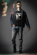 A confident man stands in a studio with cinematic lighting and analog light leaks, wearing a denim jacket over a black T-shirt that reads “PLEASE! DON’T TOUCH ME.” The shirt features a cartoon cat pushing away a hand. He’s also wearing sunglasses, jeans, and black boots, striking a bold, street-style pose.