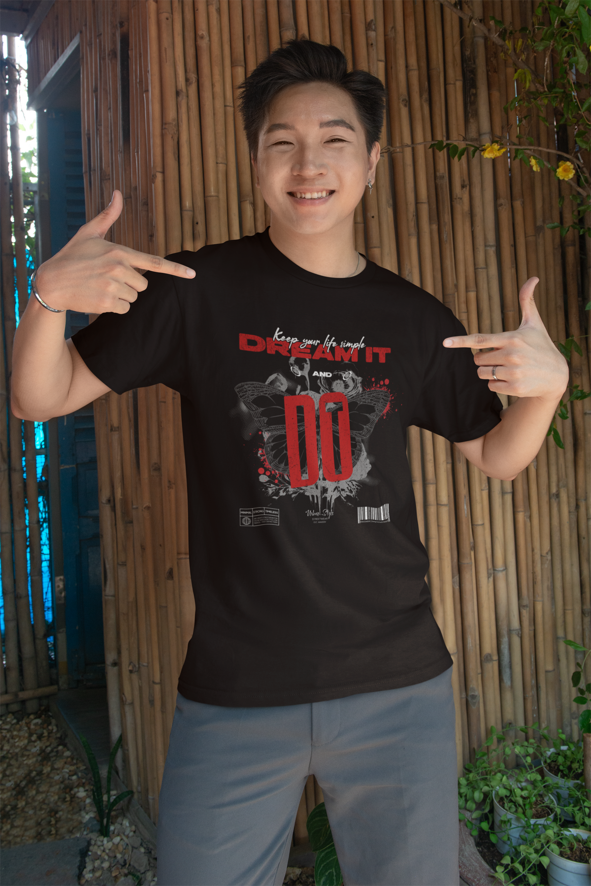 A cheerful young man stands in front of a bamboo wall, smiling and pointing at his black Krasswerk t-shirt. The design features bold red and white text reading “Keep your life simple – DREAM IT AND DO”, with two mirrored butterflies, ink splatters, and barcode elements underneath. The shirt’s modern and motivational graphic pops against the natural background.