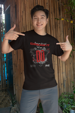 A cheerful young man stands in front of a bamboo wall, smiling and pointing at his black Krasswerk t-shirt. The design features bold red and white text reading “Keep your life simple – DREAM IT AND DO”, with two mirrored butterflies, ink splatters, and barcode elements underneath. The shirt’s modern and motivational graphic pops against the natural background.