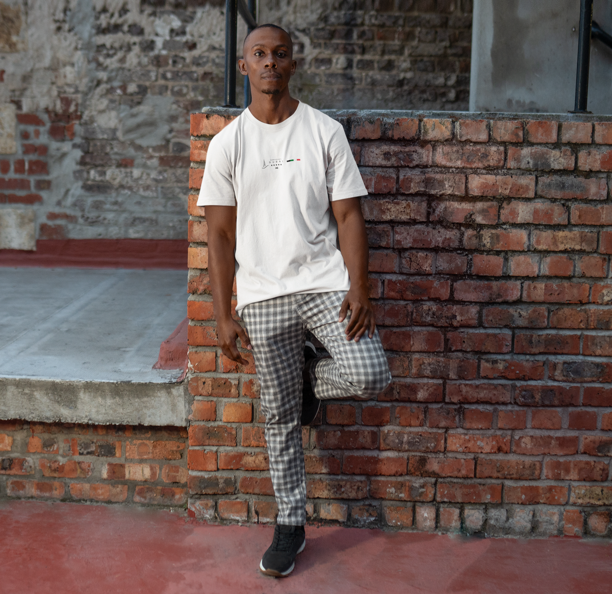 Male model leaning against a red-brick wall in a white DUBAI t-shirt, featuring a minimalist Dubai skyline and UAE flag. The scene blends urban grit with laid-back style, making the design feel grounded and cosmopolitan.