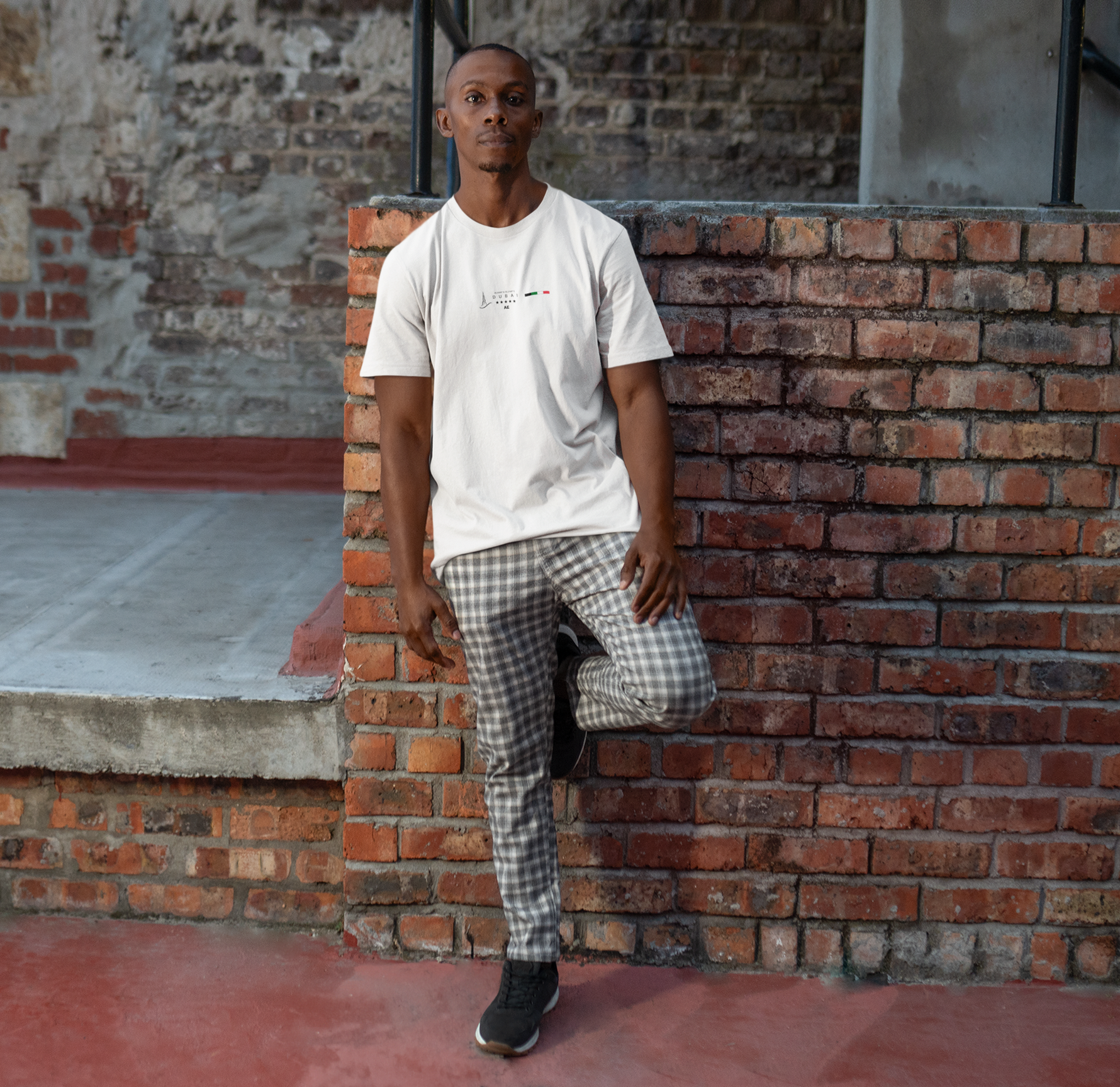 Male model leaning against a red-brick wall in a white DUBAI t-shirt, featuring a minimalist Dubai skyline and UAE flag. The scene blends urban grit with laid-back style, making the design feel grounded and cosmopolitan.