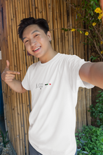 Male model smiling and pointing at the white DUBAI t-shirt in a cheerful selfie-style pose. The photo is taken outdoors in front of a bamboo wall with greenery, highlighting the shirt’s minimalist skyline and UAE flag design. The setting and natural light give it a vibrant and casual summer vibe.