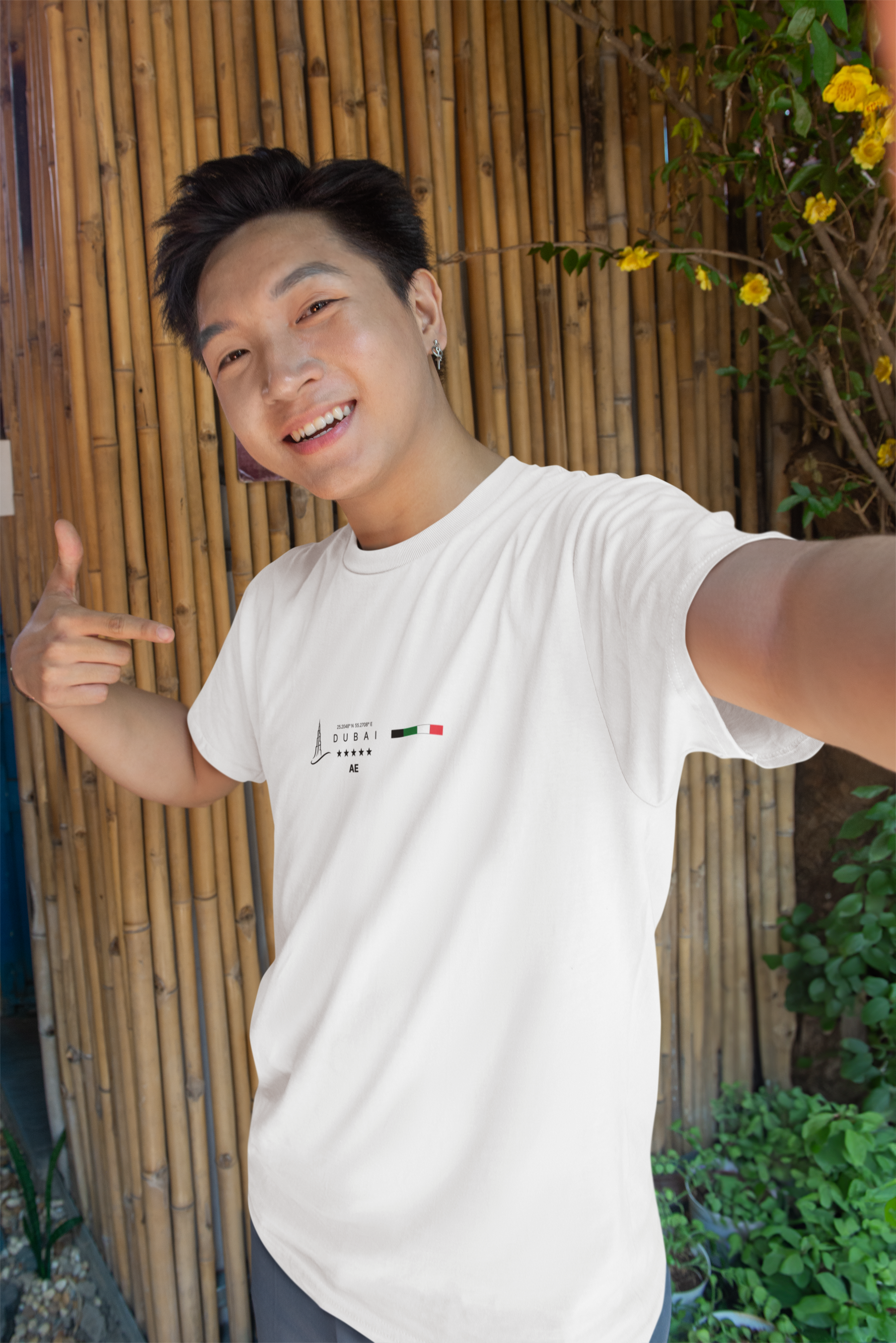 Male model smiling and pointing at the white DUBAI t-shirt in a cheerful selfie-style pose. The photo is taken outdoors in front of a bamboo wall with greenery, highlighting the shirt’s minimalist skyline and UAE flag design. The setting and natural light give it a vibrant and casual summer vibe.