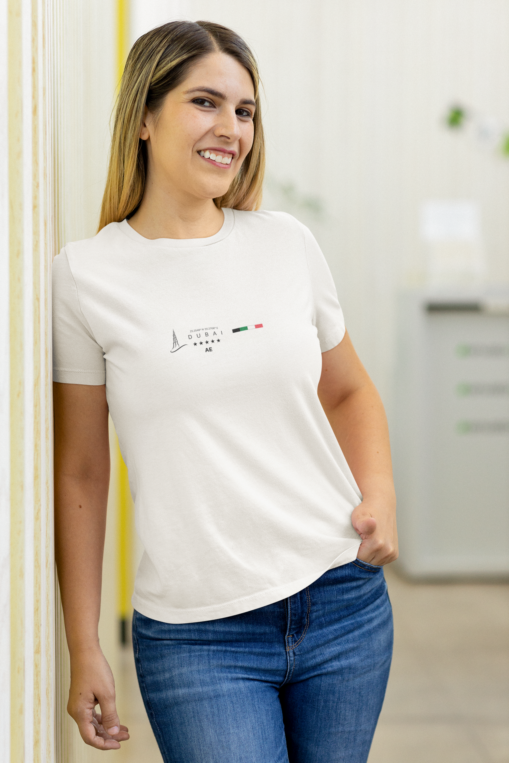 Female model in a white DUBAI t-shirt, smiling while leaning casually against a light-colored wall in an indoor office-like setting. The minimalist skyline and UAE flag design stand out against the clean background, giving the image a bright, professional tone with a relaxed feel.