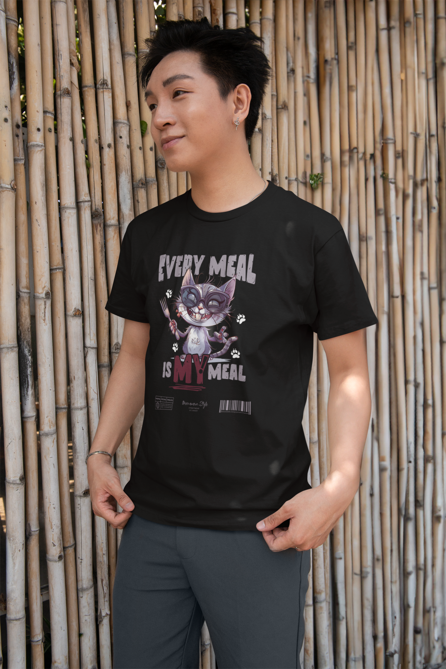 Smiling man standing in front of a bamboo wall, wearing a black “EVERY MEAL IS MY MEAL” t-shirt featuring a mischievous cat holding a fork and knife with paw prints around. The playful graphic is centered, and the setting adds a casual outdoor vibe.