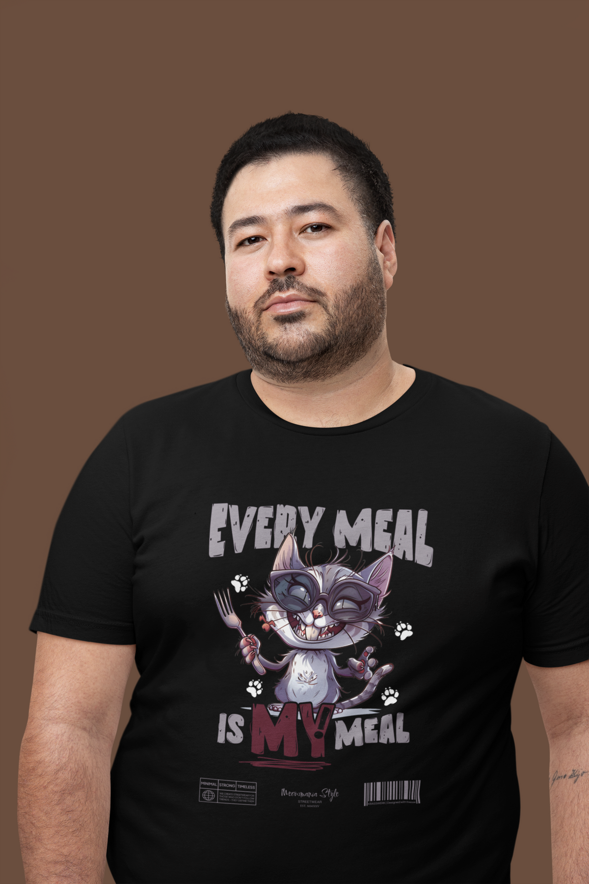 Confident man in a black “EVERY MEAL IS MY MEAL” t-shirt, featuring a mischievous cat graphic with sunglasses and a fork. Posed against a plain brown background, emphasizing the bold design and laid-back expression.