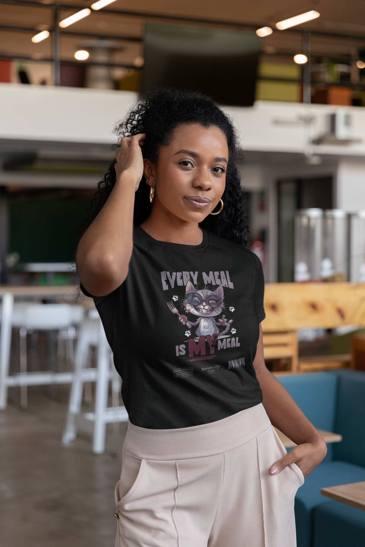 Confident woman indoors, wearing a black “EVERY MEAL IS MY MEAL” t-shirt with a grinning cat holding cutlery. The playful graphic stands out in a modern lounge-style setting, adding character to her relaxed pose and neutral-toned outfit.