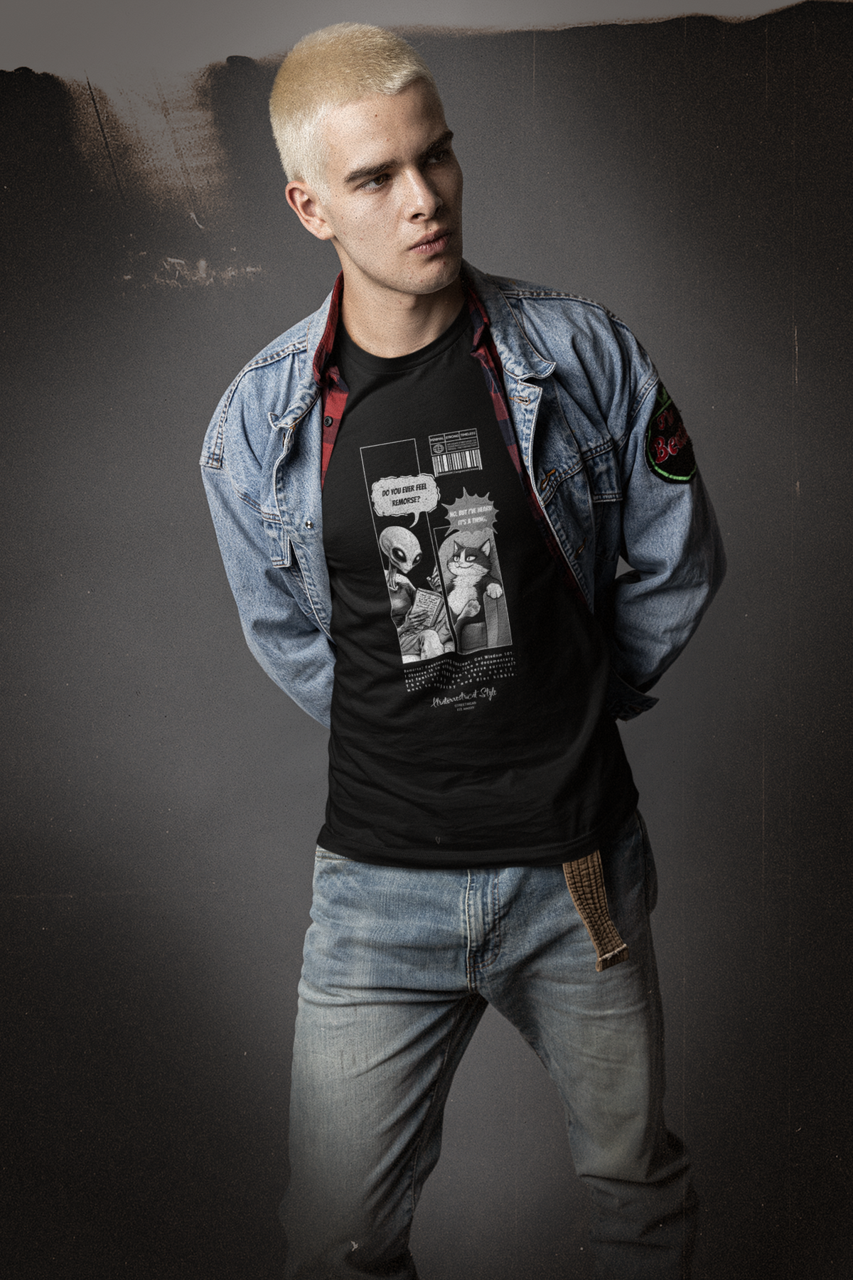 Male model with bleached hair wearing a layered denim jacket and vintage jeans, posing against a grungy studio backdrop. The black "FEEL REMORSE?" comic-style t-shirt takes center stage with a bold monochrome design: an alien holds notes while asking, “Do you ever feel remorse?” — the cat leans back, unfazed, replying, “No. But I’ve heard it’s a thing.” The shirt blends sarcastic narrative with sci-fi attitude, capturing the brand’s unapologetic edge.