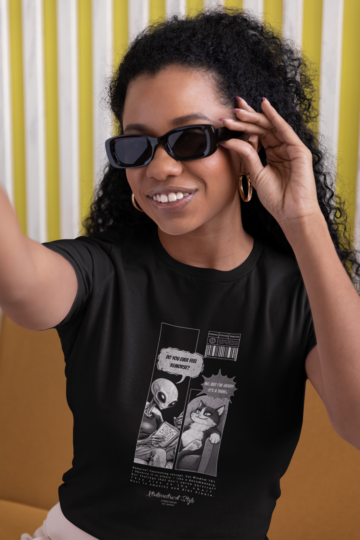 Woman model taking a cheerful selfie while adjusting her sunglasses, seated against a yellow and white background. She wears the black "FEEL REMORSE?" graphic tee, featuring a humorous monochrome comic: an alien asks, “Do you ever feel remorse?” — the cat casually replies, “No. But I’ve heard it’s a thing.” The design sits centered on the chest with barcode and microtext elements, blending sarcasm with minimalist sci-fi style.