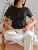 Female model sitting on a soft pink chair, wearing the FERAL t-shirt in black — calm yet confident posture highlights the claw-slashed logo and the phrase "Too Wild to Follow" on the design.