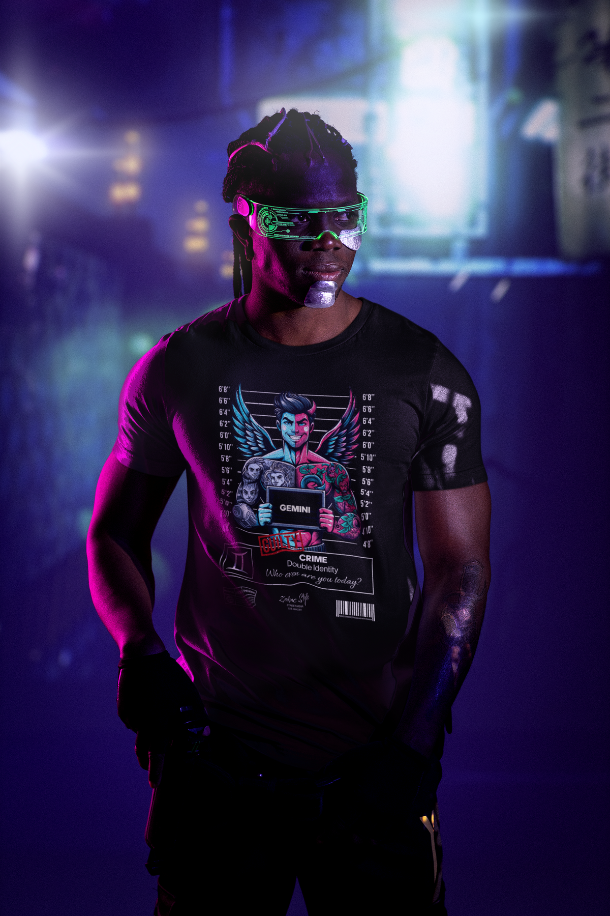 A futuristic urban scene glows in neon as the model stands confidently in the shadows, wearing the Gemini black t-shirt. The design features a winged figure split in color and personality, holding a mugshot sign — a bold visual representation of dual identity and inner contradiction.