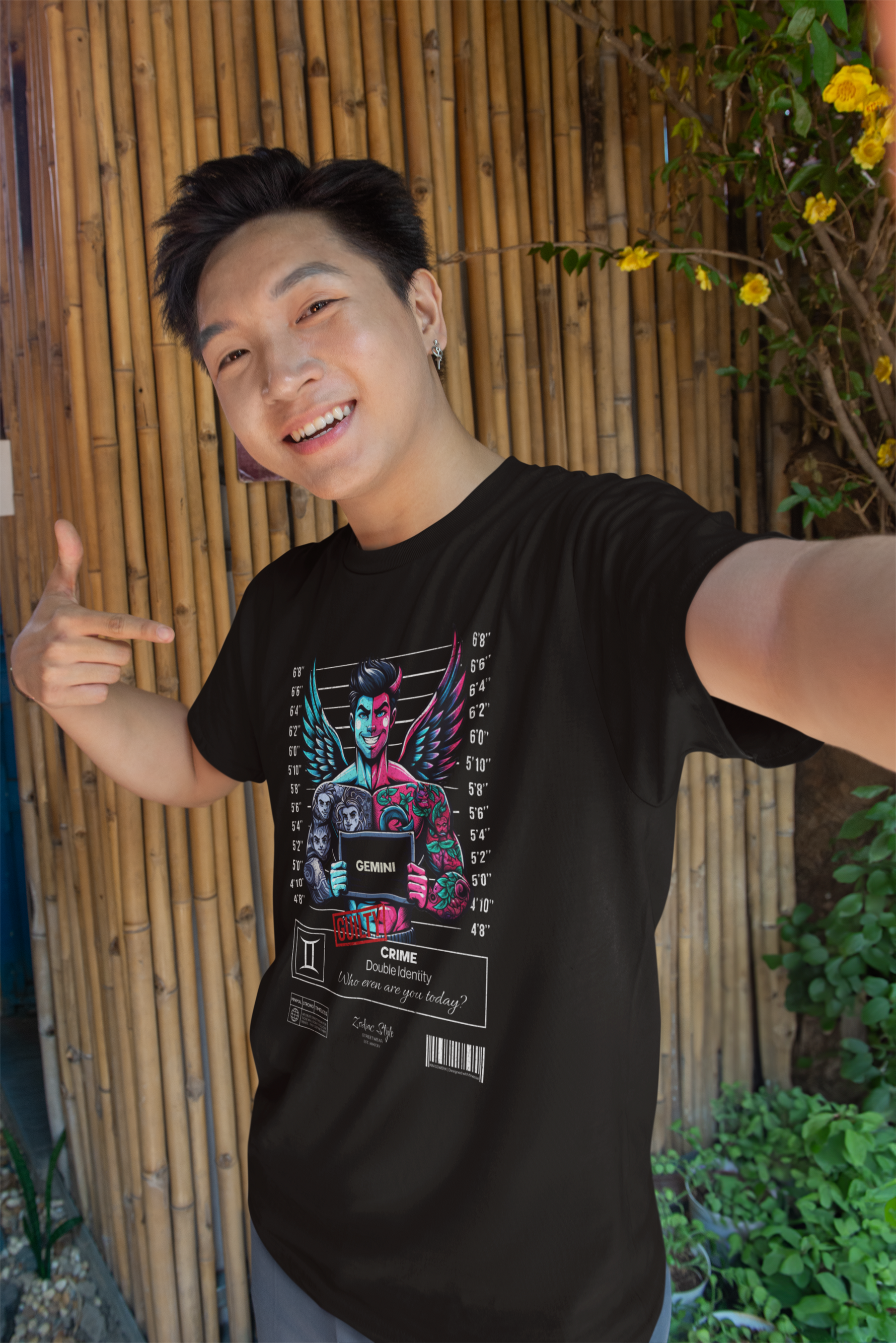 A cheerful man points proudly at his Gemini black t-shirt while standing in front of a bamboo wall. The shirt’s design shows a mischievous, double-faced figure with wings, vibrant ink, and ghostly faces on one arm — a stylish nod to the unpredictable, multi-layered nature of the Gemini spirit.