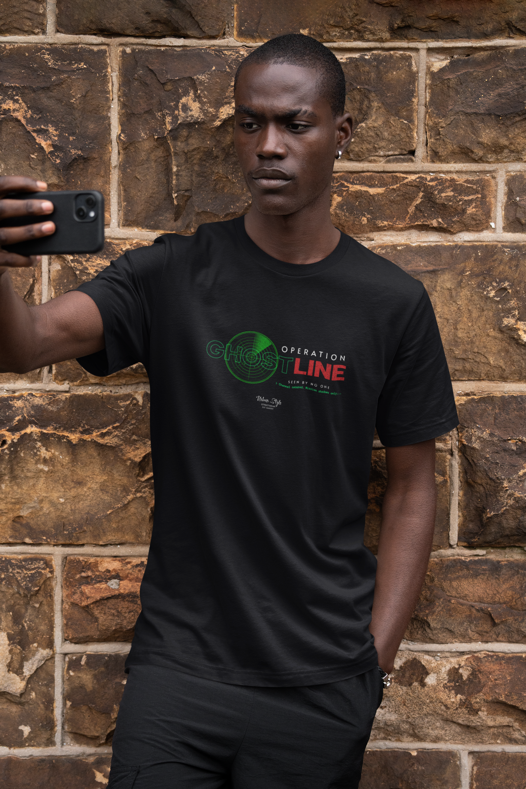 A serious-looking man in a black t-shirt stands against a rugged stone wall, holding his phone up for a selfie. The shirt reads “OPERATION GHOSTLINE” with a green radar and glitch-style red/green text, evoking a secret mission vibe. The earthy texture of the wall complements the stealthy tone of the design.