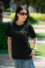 A confident woman in sunglasses poses outdoors in a black “OPERATION GHOSTLINE” t-shirt. The design features a green radar graphic and glitch-style red text. Sunlight highlights her face and shirt, while the blurred greenery and pavement in the background create a casual yet stealth-inspired city look.