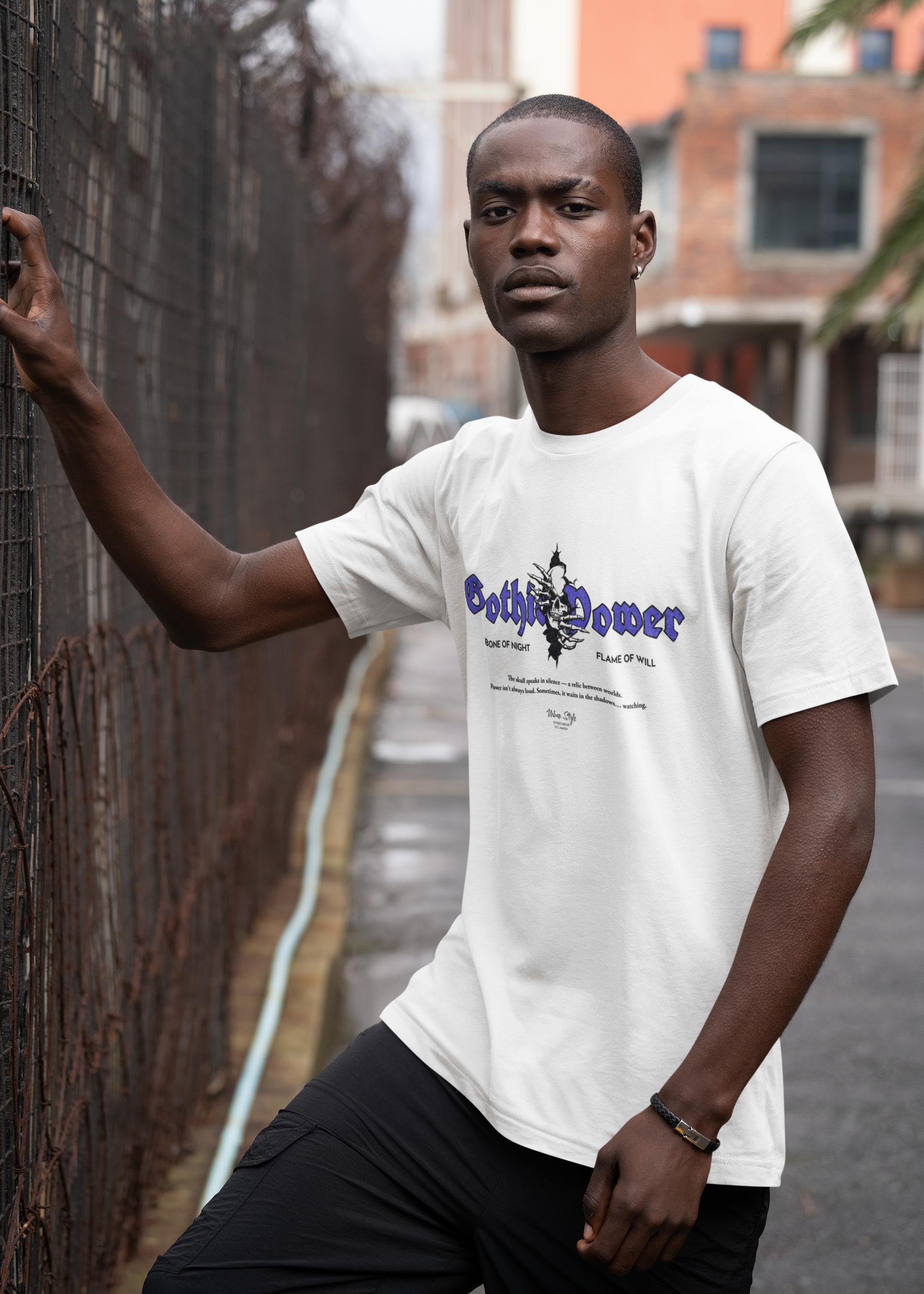 Male model in an urban alleyway, leaning on a fence while wearing the "Gothic Power" t-shirt in white. The purple gothic text and dark skull design contrast the clean fabric, evoking a modern, gritty aesthetic.