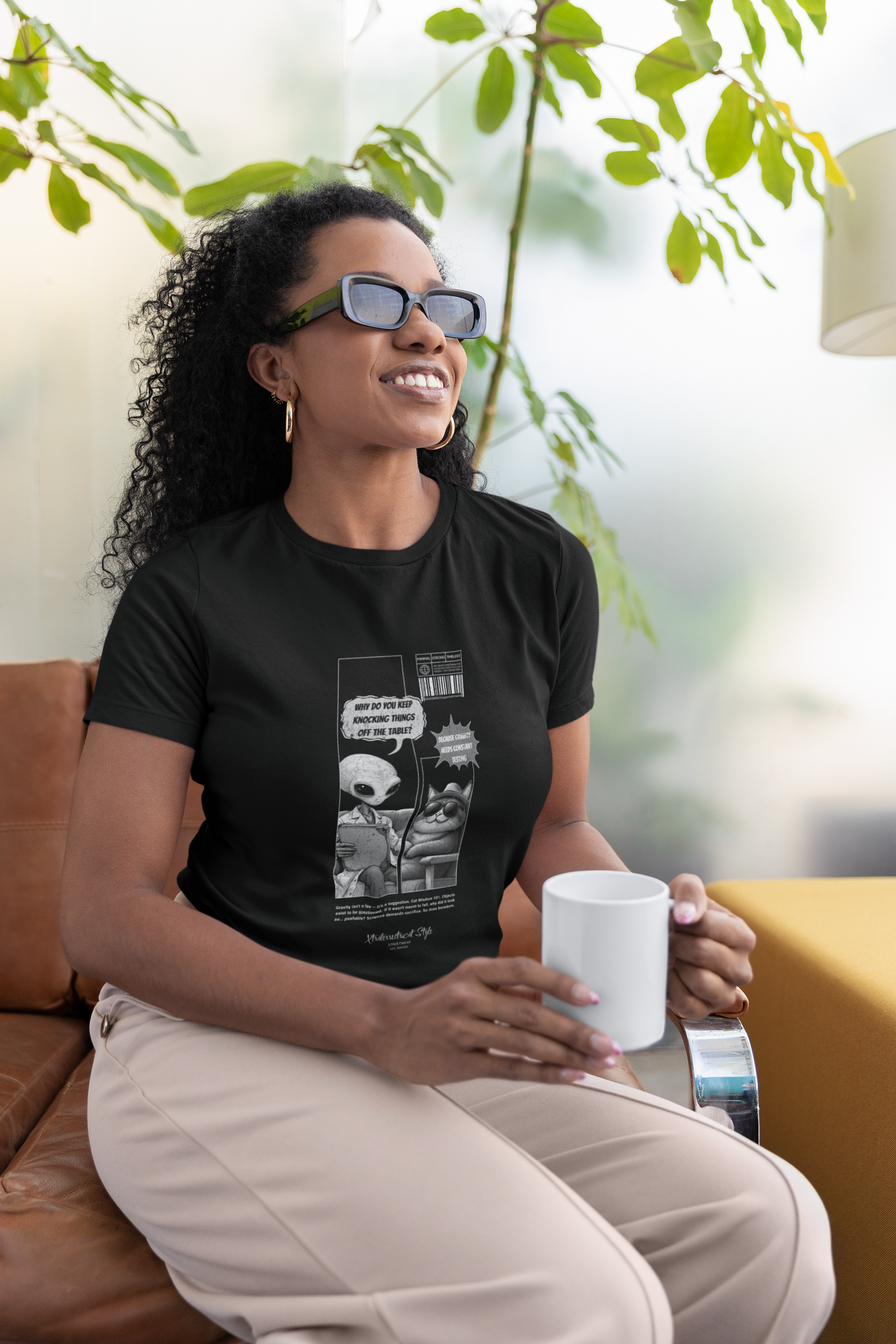 Smiling woman wearing a black t-shirt with a comic-style alien and cat illustration. The alien asks, "Why do you keep knocking things off the table?" and the cat replies, "Because gravity needs constant testing." The design features barcode graphics and fine print at the bottom, styled like a satirical scientific report. The model is seated on a modern leather chair, holding a white coffee mug in a bright indoor space filled with soft natural light and green plants in the background.