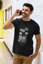Man standing in a bright hallway, smiling while talking on the phone, wearing a black t-shirt with a comic-style graphic. The design features an alien holding a clipboard asking, “Why do you keep knocking things off the table?” and a relaxed cat in sunglasses replying, “Because gravity needs constant testing.” The print includes a barcode and humorous fine print below. Casual indoor office-style setting with soft lighting.